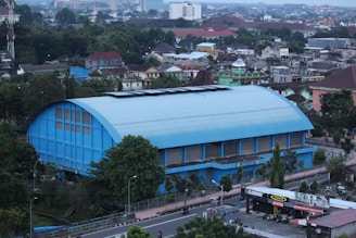 A large blue sports hall or gymnasium with an arched roof is prominently situated in the center of a bustling urban area. Surrounding the building are numerous trees and other structures, including residential and commercial buildings. The view extends to the background with dense urban development and a variety of rooftops. There are roads and some vehicles visible in the foreground.