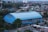 A large blue sports hall or gymnasium with an arched roof is prominently situated in the center of a bustling urban area. Surrounding the building are numerous trees and other structures, including residential and commercial buildings. The view extends to the background with dense urban development and a variety of rooftops. There are roads and some vehicles visible in the foreground.