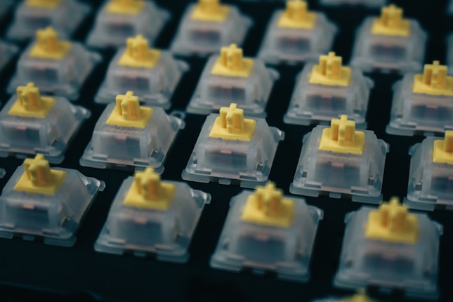 Close-up view of several mechanical keyboard switches arranged in orderly rows. Each switch is transparent with a yellow top part, set against a dark background. The focus is on the upper parts of the switches, highlighting their structure and alignment.