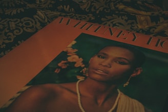 A close-up of a magazine cover featuring a woman with a serene expression. She is adorned in earrings and a pearl necklace, set against a backdrop of flowers. The cover is placed on a surface with an ornate, patterned fabric.