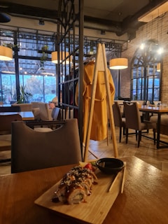 A warm and cozy restaurant interior with wooden and metal decor. A plate of sushi garnished with sauce and sesame seeds is placed on the table in the foreground. Behind it, there are tables with chairs, a coat rack with a yellow jacket, and large windows allowing natural light to illuminate the space.
