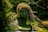 A large, intricately designed topiary sculpture of a woman's face and hand, integrated into a lush garden setting. The sculpture is covered in a variety of plants and flowers, with green, pink, and yellow tones. Water is flowing from the hand into a small pool, and the surrounding greenery is dense with various shades of foliage.