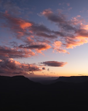 The image showcases a picturesque sunset with an array of clouds painted in soft hues of orange and pink against a backdrop of a fading blue sky. Below the sky, there is a silhouette of a mountain range, adding depth to the landscape.