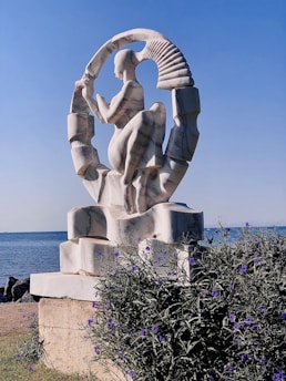 A marble sculpture of a human figure is placed on a stone pedestal near a body of water. The sculpture features a dynamic pose, with the figure surrounded by an abstract circular design. The background includes a clear blue sky, and the foreground has greenery with small purple flowers.