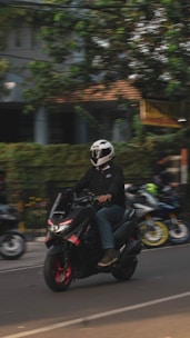 A motorcyclist wearing a black jacket and a white helmet rides a black motorcycle with red accents on a city street. The background includes blurred trees and urban buildings, suggesting motion.
