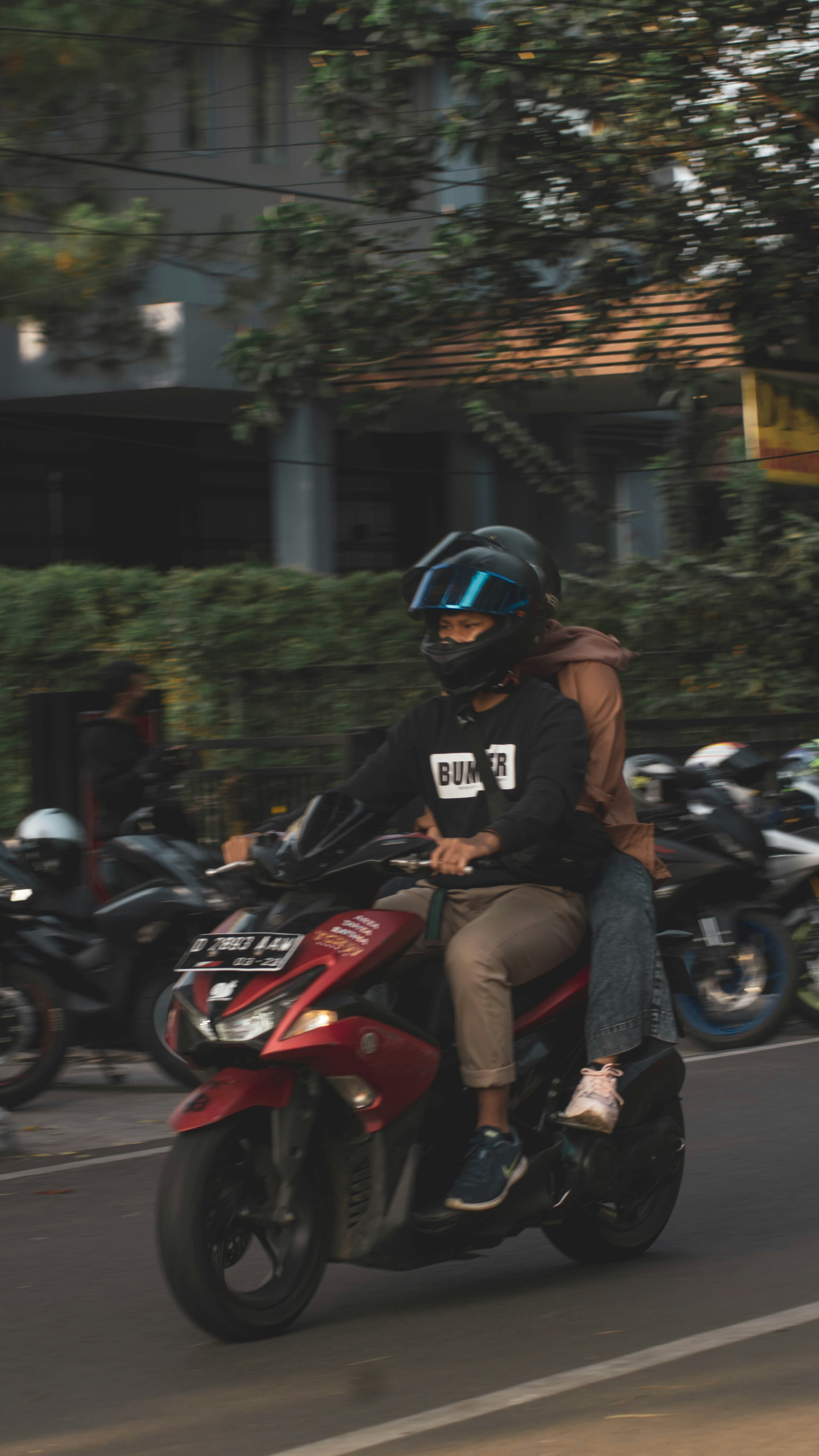 A person wearing a helmet rides a red scooter on a street, with another person sitting behind them holding on. The background features several parked motorcycles and a building with green foliage.