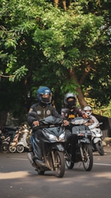 Two motorcyclists wearing helmets are riding their bikes on a road surrounded by lush green trees. Both are dressed in protective gear, and several other parked motorcycles can be seen in the background.