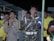A group of people, predominantly men, are engaged in a moment of prayer or reflection. One individual wears a camouflage uniform with a white belt, raising both hands, conveying a sense of devotion. Others are dressed in yellow jackets and traditional hats. Their expressions range from focused to emotional, with hands clasped or raised. The setting has a rustic backdrop with wooden and corrugated metal elements.