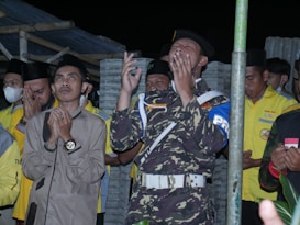 A group of people, predominantly men, are engaged in a moment of prayer or reflection. One individual wears a camouflage uniform with a white belt, raising both hands, conveying a sense of devotion. Others are dressed in yellow jackets and traditional hats. Their expressions range from focused to emotional, with hands clasped or raised. The setting has a rustic backdrop with wooden and corrugated metal elements.