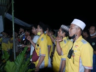 A group of men stand closely together, some wearing yellow uniforms with patches and others in casual attire. They appear to be raising their fists in a gesture, possibly symbolizing unity or solidarity. One man in the foreground has a walkie-talkie attached to his shirt and wears a white cap. The scene is outdoors with green plants visible at the bottom of the image.