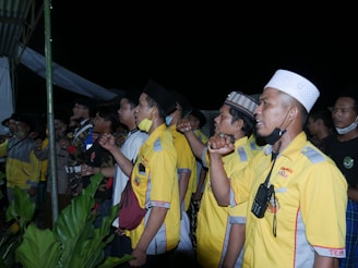 A group of men stand closely together, some wearing yellow uniforms with patches and others in casual attire. They appear to be raising their fists in a gesture, possibly symbolizing unity or solidarity. One man in the foreground has a walkie-talkie attached to his shirt and wears a white cap. The scene is outdoors with green plants visible at the bottom of the image.