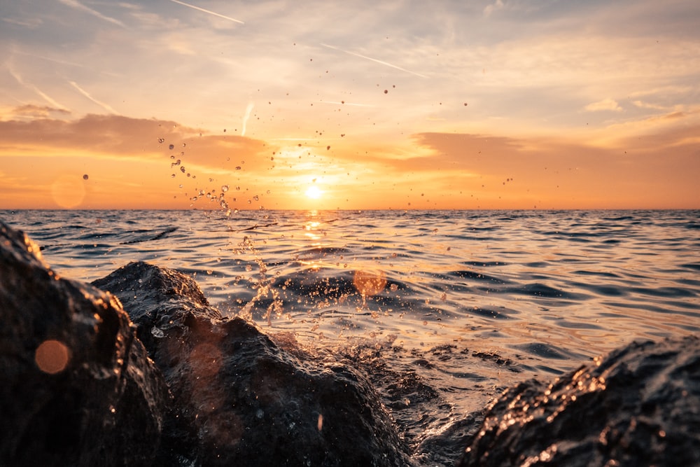 the sun is setting over the ocean with rocks in the foreground