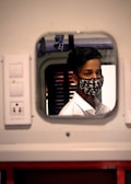 A young individual wearing a face mask with a star pattern, reflecting in a small, rectangular mirror. The person is dressed in a white shirt. The setting appears to be indoors with some electrical outlets visible on the wall near the mirror. The background includes a window with horizontal slats.