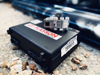 A black plastic case labeled with 'PLASSON' rests on a rough, stony surface. On top of the case, a small metallic mechanical part with several cylindrical protrusions is placed. The surrounding area includes scattered small rocks and a blurred background of pipes or similar industrial materials.