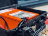 A close-up view of a hard plastic case with a latch open displaying an electronic device inside. The device is orange with a number of controls and indicators visible. Red and black cables or connectors are visible in the foreground, indicating potential use in electrical or technical applications.