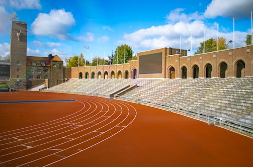 An outdoor athletic stadium featuring a red running track with lanes marked in white curves around the edge of the field. The seating area consists of stone bleachers that rise to meet an old brick building with arched windows and a scoreboard. Trees with autumn foliage can be seen in the background under a bright blue sky with scattered clouds.