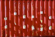 Several tin cans are suspended with colorful strings against a red corrugated metal wall. Each can contains small green plants, creating a vertical garden.