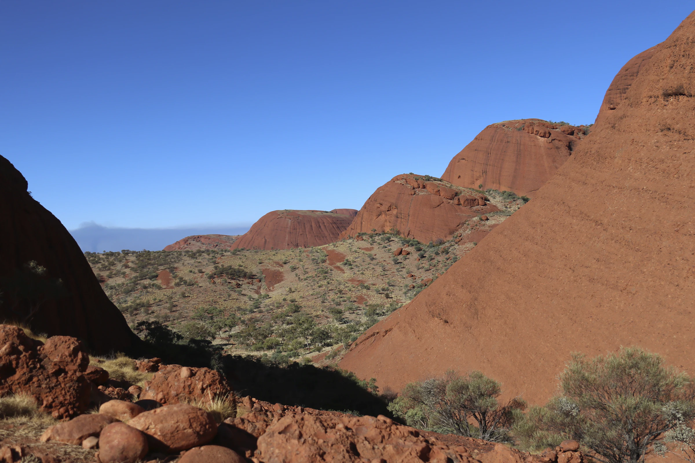 View from a rocky slope across layered red-rock hills and rounded formations, with sparse shrubs beneath a clear blue sky.