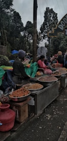 A bustling outdoor market scene with vendors selling various goods. Large bowls filled with different kinds of snacks or grains are prominently displayed in the foreground. People, dressed in warm clothing, are either sitting or standing around the market. Tall trees and an overcast sky create a natural backdrop.