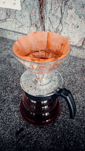 A pour-over coffee dripper with a used paper filter sits atop a glass carafe. The dripper is made of clear glass with coffee grounds visible in the filter. The carafe below contains freshly brewed coffee. The setting appears to be a granite countertop, with a wall outlet partially visible in the background.