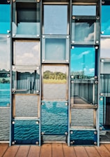 A series of glass panels arranged in a grid pattern, reflecting a scenic view of a serene landscape with greenery and a body of water. The panels are mounted using metallic bolts, creating a structured and geometric appearance. The view through the glass includes a clear blue sky with some clouds.