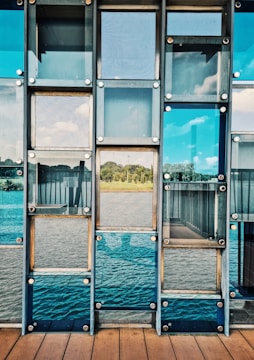 A series of glass panels arranged in a grid pattern, reflecting a scenic view of a serene landscape with greenery and a body of water. The panels are mounted using metallic bolts, creating a structured and geometric appearance. The view through the glass includes a clear blue sky with some clouds.