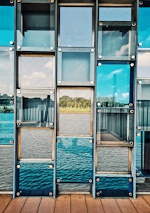 A series of glass panels arranged in a grid pattern, reflecting a scenic view of a serene landscape with greenery and a body of water. The panels are mounted using metallic bolts, creating a structured and geometric appearance. The view through the glass includes a clear blue sky with some clouds.