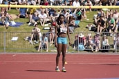 An athlete stands on a red running track, wearing a sports bra and shorts with a number bib. A yellow bar is positioned above as part of a high jump setup. In the background, a crowd sits on a grassy area, some on blankets, watching the event on a sunny day.