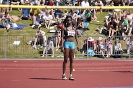 An athlete stands on a red running track, wearing a sports bra and shorts with a number bib. A yellow bar is positioned above as part of a high jump setup. In the background, a crowd sits on a grassy area, some on blankets, watching the event on a sunny day.