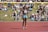 An athlete stands on a red running track, wearing a sports bra and shorts with a number bib. A yellow bar is positioned above as part of a high jump setup. In the background, a crowd sits on a grassy area, some on blankets, watching the event on a sunny day.