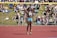 An athlete stands on a red running track, wearing a sports bra and shorts with a number bib. A yellow bar is positioned above as part of a high jump setup. In the background, a crowd sits on a grassy area, some on blankets, watching the event on a sunny day.