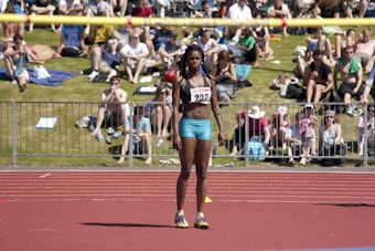 An athlete stands on a red running track, wearing a sports bra and shorts with a number bib. A yellow bar is positioned above as part of a high jump setup. In the background, a crowd sits on a grassy area, some on blankets, watching the event on a sunny day.