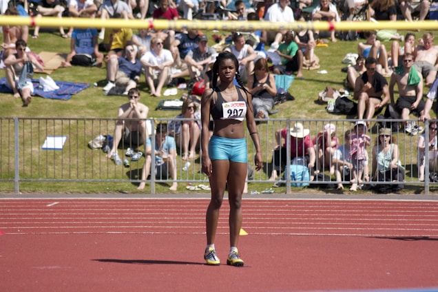 An athlete stands on a red running track, wearing a sports bra and shorts with a number bib. A yellow bar is positioned above as part of a high jump setup. In the background, a crowd sits on a grassy area, some on blankets, watching the event on a sunny day.