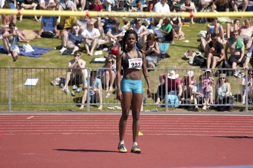 An athlete stands on a red running track, wearing a sports bra and shorts with a number bib. A yellow bar is positioned above as part of a high jump setup. In the background, a crowd sits on a grassy area, some on blankets, watching the event on a sunny day.