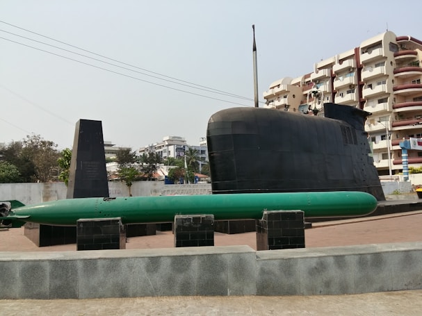 A large black submarine is displayed outdoors on a raised platform, accompanied by a green torpedo positioned on the ground in front. The background features residential buildings, trees, and a clear sky, indicating an urban setting.