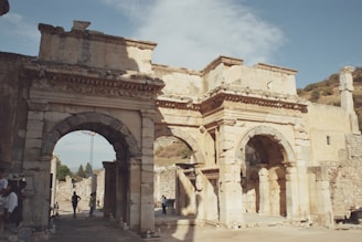 An ancient stone structure features two large arched gateways and intricate carvings on the facade. The architecture showcases historical significance, with its weathered stone and surrounding ruins. People are seen walking through the arches, adding perspective to the grandeur of the site. The sky is partly cloudy, providing a serene backdrop.