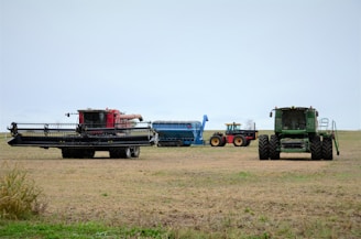 Several pieces of large agricultural machinery are positioned in an open field, including a red combine harvester on the left, a blue grain cart in the center, and a green combine harvester on the right. The field appears to be barren, likely post-harvest, with a cloudy sky overhead.