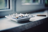 Numerous cigarette butts are scattered in a white ashtray on a windowsill, with a single cigarette resting beside it. The setting appears to be indoors, with soft, natural light filtering through a window.
