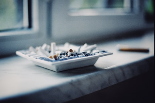 Numerous cigarette butts are scattered in a white ashtray on a windowsill, with a single cigarette resting beside it. The setting appears to be indoors, with soft, natural light filtering through a window.