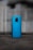 A smartphone with a glossy blue back cover is standing upright on a darker surface. The brand name 'Redmi' is visible on the lower section of the phone. The background is blurred and shows indistinct, muted colors, likely an outdoor setting.