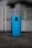 A smartphone with a glossy blue back cover is standing upright on a darker surface. The brand name 'Redmi' is visible on the lower section of the phone. The background is blurred and shows indistinct, muted colors, likely an outdoor setting.
