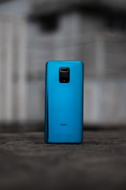 A smartphone with a glossy blue back cover is standing upright on a darker surface. The brand name 'Redmi' is visible on the lower section of the phone. The background is blurred and shows indistinct, muted colors, likely an outdoor setting.