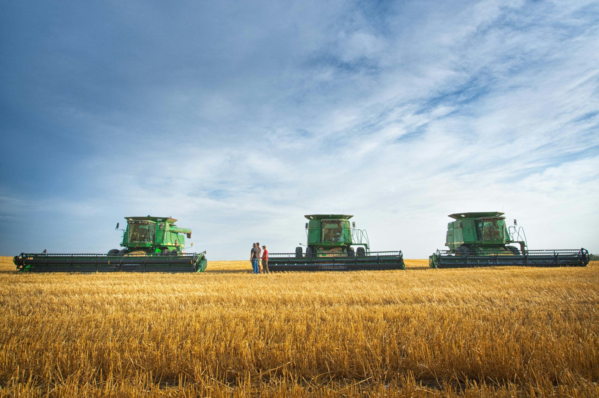 Front view of line of John Deere combine harvesters farm workers in wheat field blue sky clouds wheat stubble grain