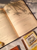Open book with Arabic text, placed on a white surface with sunlight casting shadows from a nearby plant. A smaller booklet with a yellow and brown cover is partially visible at the bottom right.