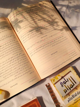 Open book with Arabic text, placed on a white surface with sunlight casting shadows from a nearby plant. A smaller booklet with a yellow and brown cover is partially visible at the bottom right.