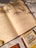 Open book with Arabic text, placed on a white surface with sunlight casting shadows from a nearby plant. A smaller booklet with a yellow and brown cover is partially visible at the bottom right.
