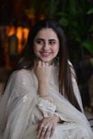 A woman in an elegant, traditional light-colored outfit, adorned with intricate lace and embroidery. She is smiling and resting her chin on her hand, wearing silver jewelry including a ring and bracelets. The background contains blurred greenery and warm lighting.