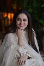 A woman in an elegant, traditional light-colored outfit, adorned with intricate lace and embroidery. She is smiling and resting her chin on her hand, wearing silver jewelry including a ring and bracelets. The background contains blurred greenery and warm lighting.