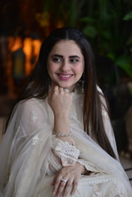 A woman in an elegant, traditional light-colored outfit, adorned with intricate lace and embroidery. She is smiling and resting her chin on her hand, wearing silver jewelry including a ring and bracelets. The background contains blurred greenery and warm lighting.