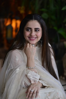 A woman in an elegant, traditional light-colored outfit, adorned with intricate lace and embroidery. She is smiling and resting her chin on her hand, wearing silver jewelry including a ring and bracelets. The background contains blurred greenery and warm lighting.